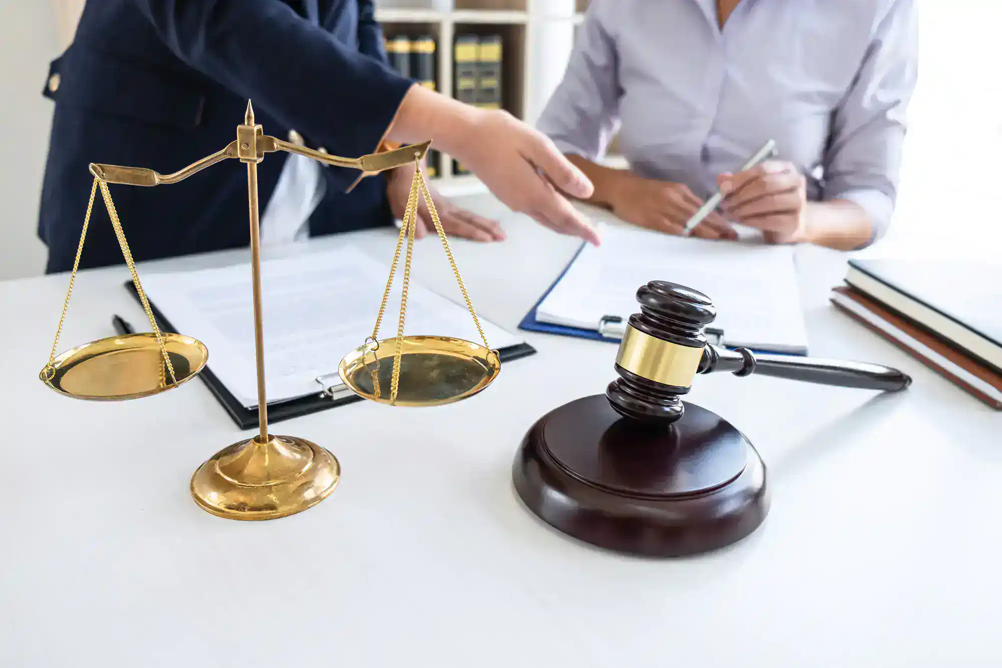Two people sit at a desk with legal documents, a golden balance scale, and a wooden judge&rsquo;s gavel in the foreground, symbolizing law and justice. One person gestures while the other writes on a clipboard.