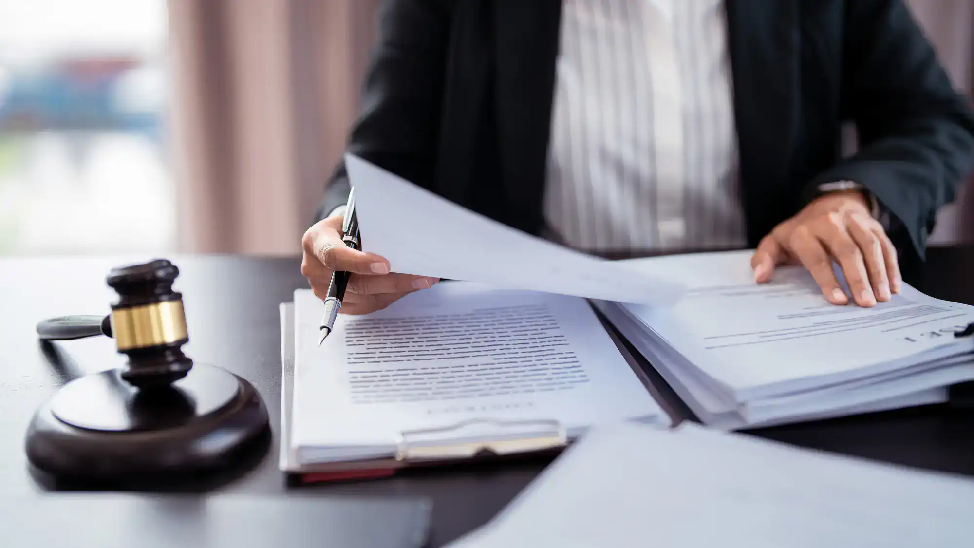 A person in business attire reviews legal documents at a desk, with a gavel nearby and stacks of paperwork, suggesting a legal or courtroom setting.