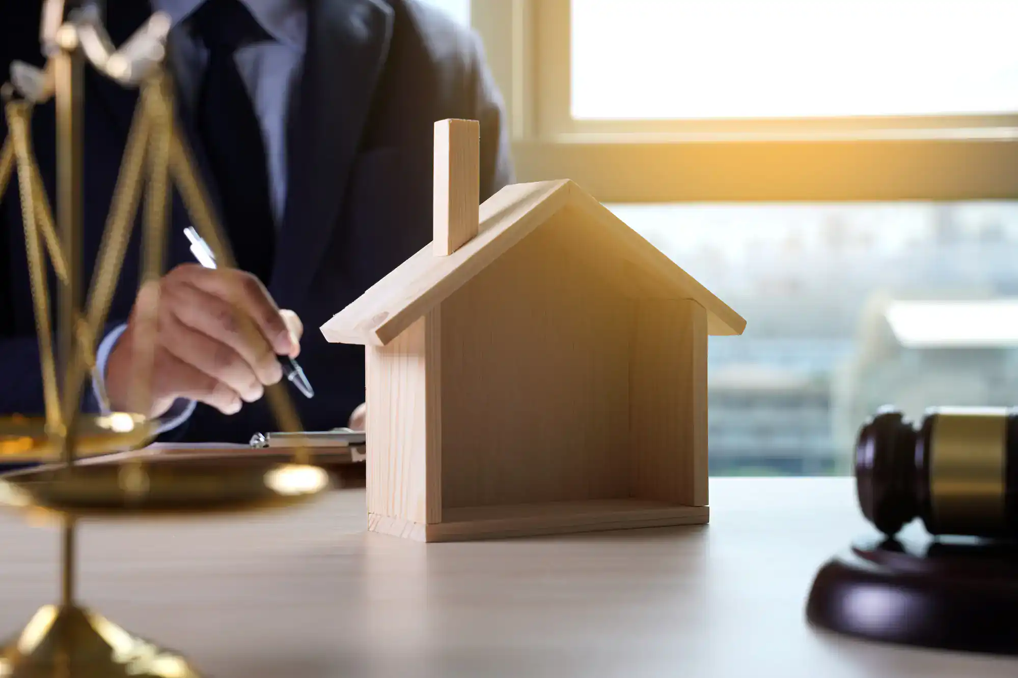 A person in a suit writes on paper at a desk with a wooden house model, a gavel, and a set of scales, symbolizing legal or real estate matters. Sunlight shines through a window in the background.