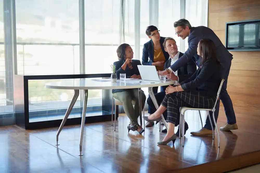 Five business professionals gather around a table in a modern office, discussing ideas and looking at a laptop. Sunlight streams through large windows, creating a collaborative atmosphere.